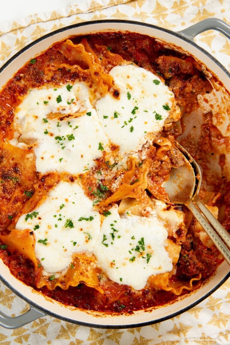A close-up of a skillet lasagna topped with melted cheese and sprinkled with herbs, with a serving spoon partially inside the dish.