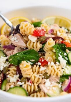 A close-up of a Greek tuna pasta salad with spinach, feta cheese, cucumbers, red onions, capers, and roasted red peppers in a white bowl, garnished with lemon slices.