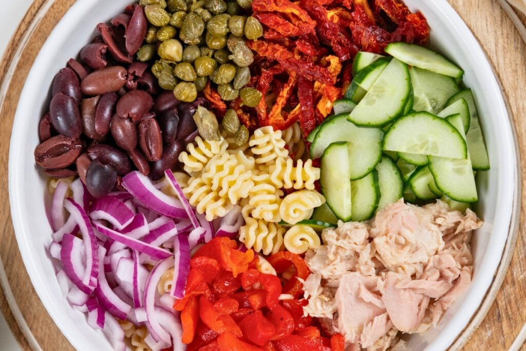 Greek Tuna Pasta Salad has salty olives, cool cucumber, and creamy feta in a lemon garlic dressing. No mayo, no oven, ready in 30 minutes! A bowl filled with sliced cucumbers, sun-dried tomatoes, capers, Kalamata olives, cooked pasta, tuna, red onions, and red bell peppers, all arranged in separate sections.