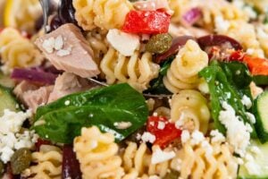 A close-up of a Greek tuna pasta salad with spinach, feta cheese, cucumbers, red onions, capers, and roasted red peppers in a white bowl, garnished with lemon slices.