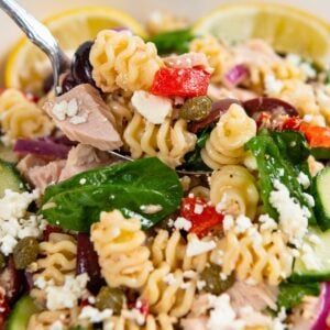 A close-up of a Greek tuna pasta salad with spinach, feta cheese, cucumbers, red onions, capers, and roasted red peppers in a white bowl, garnished with lemon slices.