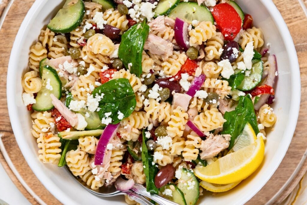 Greek Tuna Pasta Salad has salty olives, cool cucumber, and creamy feta in a lemon garlic dressing. No mayo, no oven, ready in 30 minutes! A bowl of tuna pasta salad with rotini, spinach, cucumbers, red onions, olives, capers, feta cheese, and lemon wedges, with serve spoons tucked in the bowl on the side.