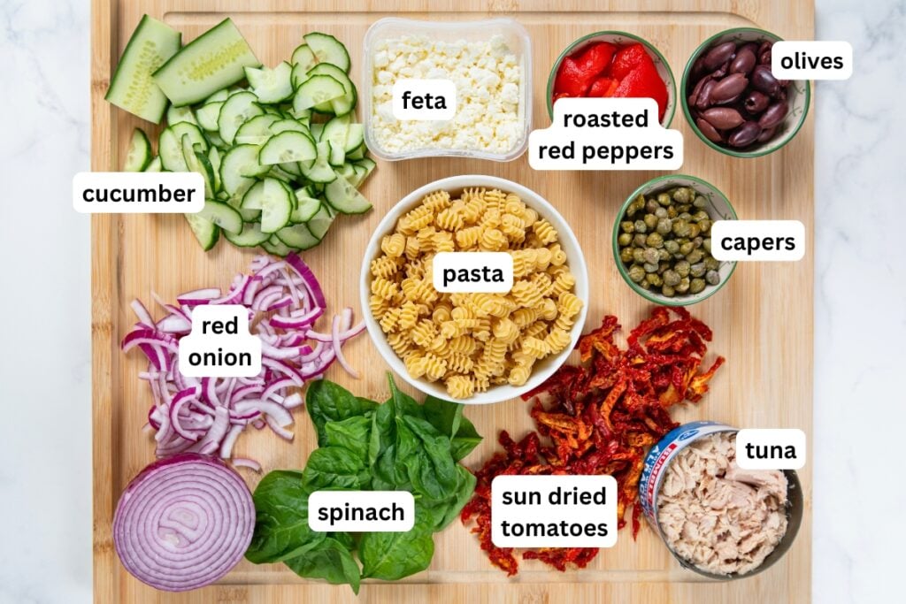Greek Tuna Pasta Salad has salty olives, cool cucumber, and creamy feta in a lemon garlic dressing. No mayo, no oven, ready in 30 minutes! A wooden board with labeled ingredients: cucumber, feta, roasted red peppers, olives, capers, tuna, sun dried tomatoes, spinach, red onion, and cooked pasta, all arranged in bowls or piles.