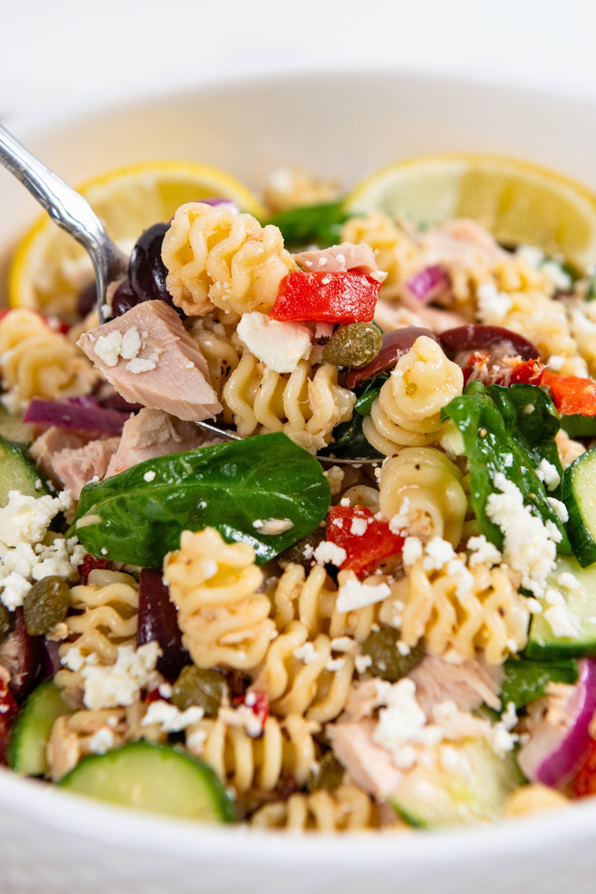 Greek Tuna Pasta Salad has salty olives, cool cucumber, and creamy feta in a lemon garlic dressing. No mayo, no oven, ready in 30 minutes! A close-up of a Greek tuna pasta salad with spinach, feta cheese, cucumbers, red onions, capers, and roasted red peppers in a white bowl, garnished with lemon slices.