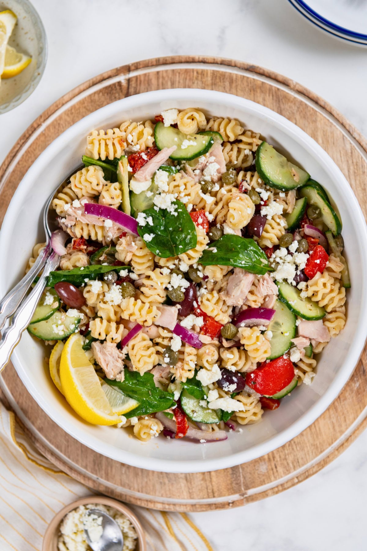 Greek Tuna Pasta Salad has salty olives, cool cucumber, and creamy feta in a lemon garlic dressing. No mayo, no oven, ready in 30 minutes! A bowl of Greek tuna pasta salad with spiral noodles, spinach, cucumbers, red onions, tomatoes, feta cheese, and tuna, garnished with lemon wedges.