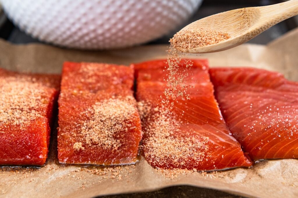 Sweet, savory, and a little smoky, this Pan Seared Salmon is moist and flaky with a caramelized brown sugar crust. Ready in just 15 minutes! A wooden spoon sprinkles a spiced brown sugar rub onto three raw salmon fillets arranged on parchment paper.