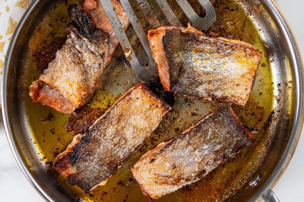 Sweet, savory, and a little smoky, this Pan Seared Salmon is moist and flaky with a caramelized brown sugar crust. Ready in just 15 minutes! Four pieces of pan seared salmon with crispy skin are frying in a large metal pan with oil. A metal spatula rests in the pan, partially under one of the fish pieces.