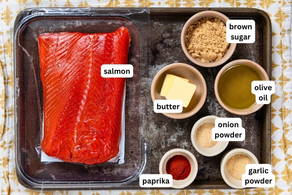 Sweet, savory, and a little smoky, this Pan Seared Salmon is moist and flaky with a caramelized brown sugar crust. Ready in just 15 minutes! Raw salmon fillet on a tray with labeled bowls of brown sugar, olive oil, butter, onion powder, garlic powder, and paprika on a patterned surface.
