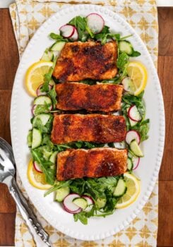 Pan seared salmon with a brown sugar crust on a bed of arugula, cucumber, and radish salad, garnished with lemon slices, served on a white oval platter with serving utensils beside it.