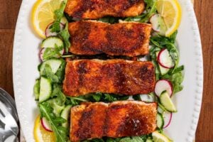 Pan seared salmon with a brown sugar crust on a bed of arugula, cucumber, and radish salad, garnished with lemon slices, served on a white oval platter with serving utensils beside it.