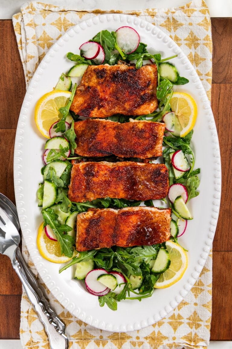 Pan seared salmon with a brown sugar crust on a bed of arugula, cucumber, and radish salad, garnished with lemon slices, served on a white oval platter with serving utensils beside it.