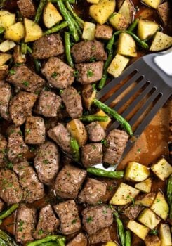 A spatula lifts seasoned and roasted steak bites and potatoes with green beans from a sheet pan.