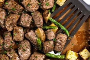 A spatula lifts seasoned and roasted steak bites and potatoes with green beans from a sheet pan.