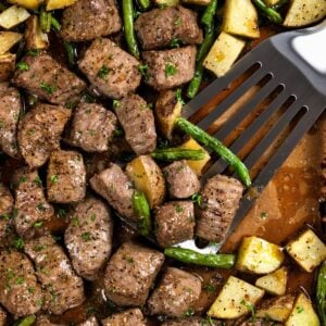 A spatula lifts seasoned and roasted steak bites and potatoes with green beans from a sheet pan.