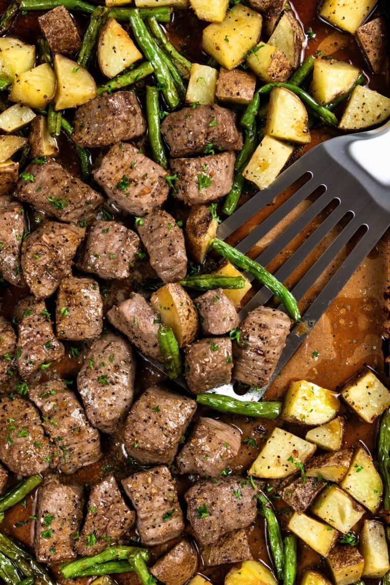 A spatula lifts seasoned and roasted steak bites and potatoes with green beans from a sheet pan.