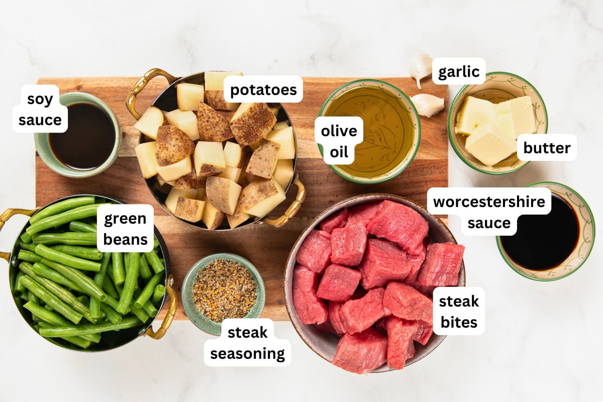 Sheet Pan Steak Bites and Potatoes gives you juicy sirloin, crispy potatoes, and roasted green beans off one pan. Ready in about 40 minutes! A wooden board with bowls of soy sauce, green beans, steak seasoning, diced potatoes, olive oil, garlic, butter, Worcestershire sauce, and raw steak bites, each item labeled.