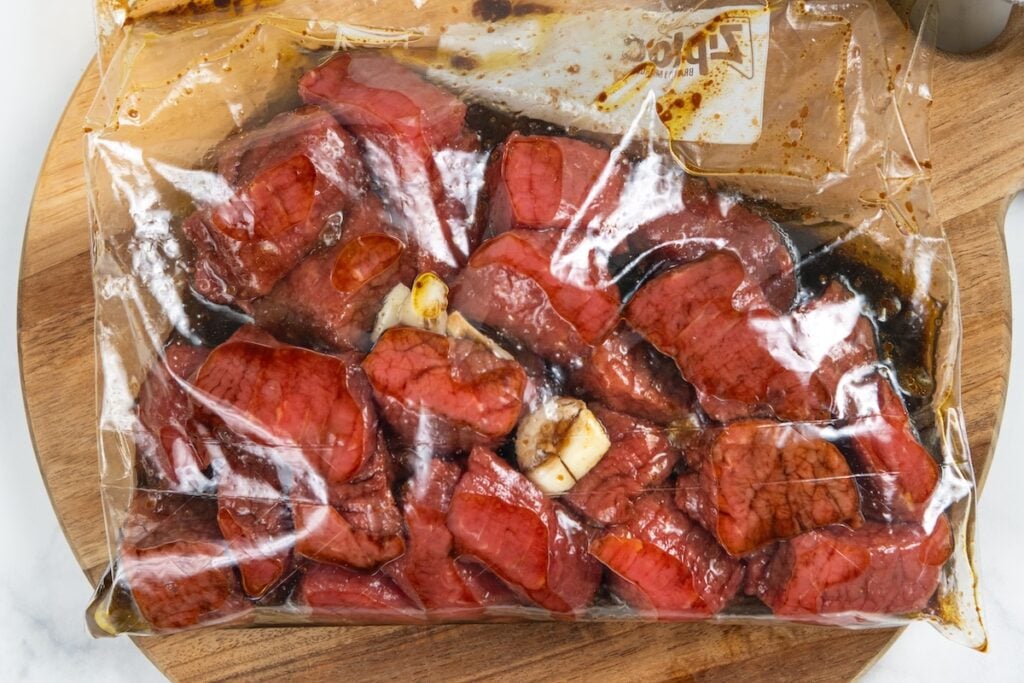 Sheet Pan Steak Bites and Potatoes gives you juicy sirloin, crispy potatoes, and roasted green beans off one pan. Ready in about 40 minutes! Raw sirloin steak bites marinating in a clear plastic zip-top bag with a dark marinade and crushed garlic cloves, placed on a wooden cutting board.