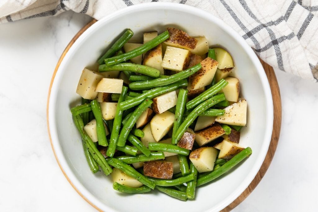 Sheet Pan Steak Bites and Potatoes gives you juicy sirloin, crispy potatoes, and roasted green beans off one pan. Ready in about 40 minutes! Raw green beans and cubed potatoes coated in oil and seasonings in a large white bowl.