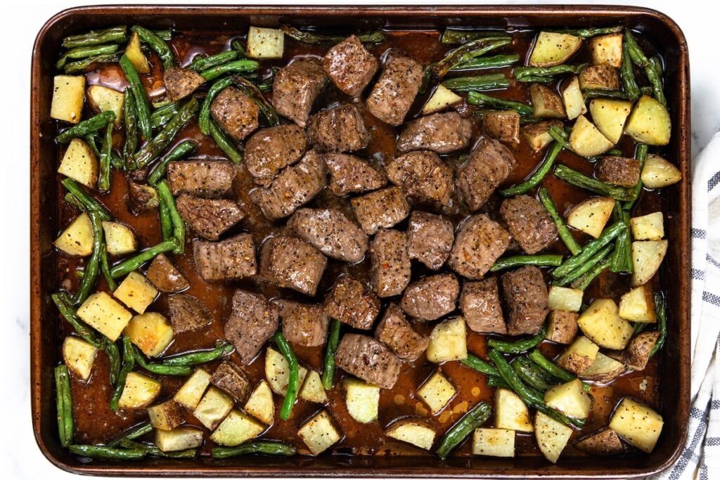 Sheet Pan Steak Bites and Potatoes gives you juicy sirloin, crispy potatoes, and roasted green beans off one pan. Ready in about 40 minutes! Sheet pan with seasoned steak bites and potatoes with green beans, all roasted and spread evenly, with some juices visible on the pan.