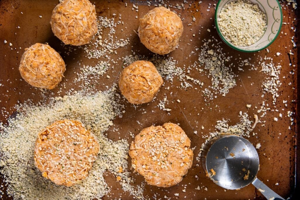 This Chicken Patty Sandwich turns two cans of chicken into a quick and easy dinner for six! Served with a tangy sauce and all the toppings. Six round, uncooked canned chicken patties made from shredded ingredients are on a brown baking sheet, surrounded by scattered breadcrumbs. A bowl with breadcrumbs and a metal measuring cup are nearby.