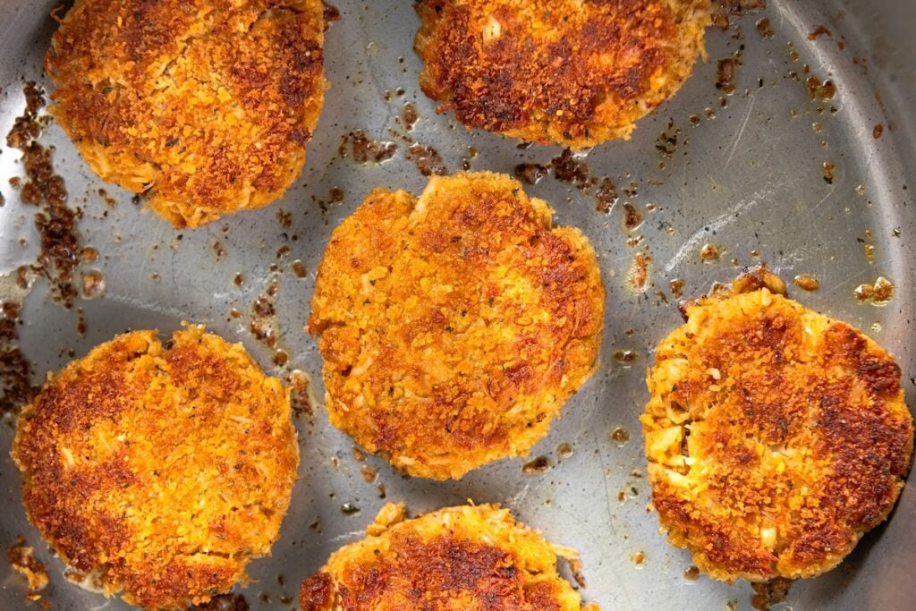 This Chicken Patty Sandwich turns two cans of chicken into a quick and easy dinner for six! Served with a tangy sauce and all the toppings. Six golden-brown patties fry in a silver pan. The patties have a crispy, textured surface and are surrounded by small crumbs.