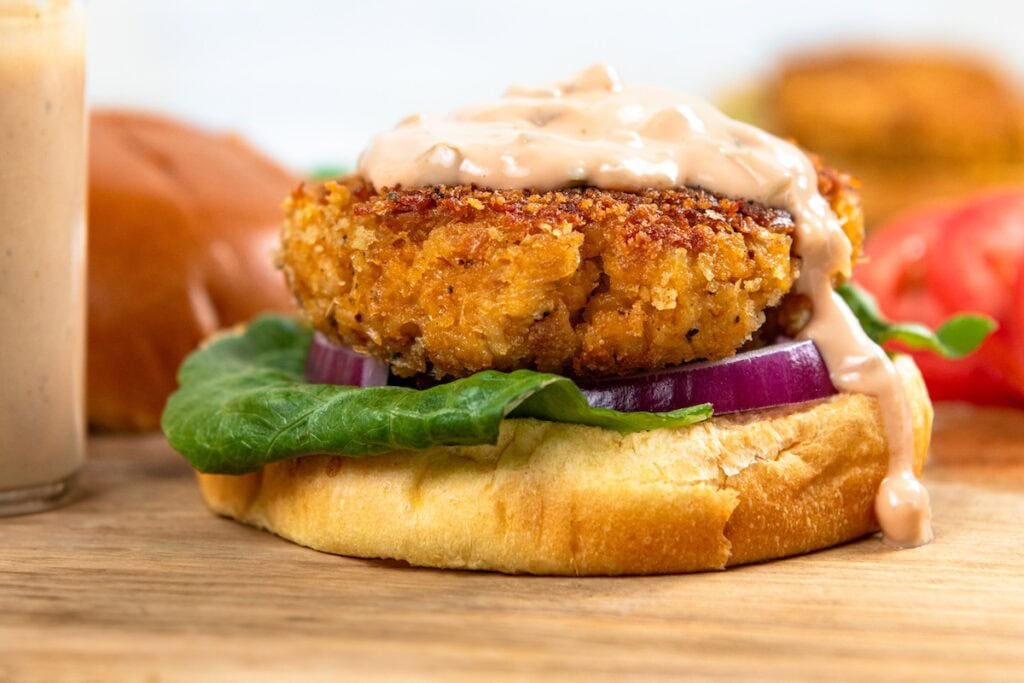 This Chicken Patty Sandwich turns two cans of chicken into a quick and easy dinner for six! Served with a tangy sauce and all the toppings. A crispy breaded patty on a bun with lettuce, red onion, and creamy sauce, with tomato and bun halves in the background.