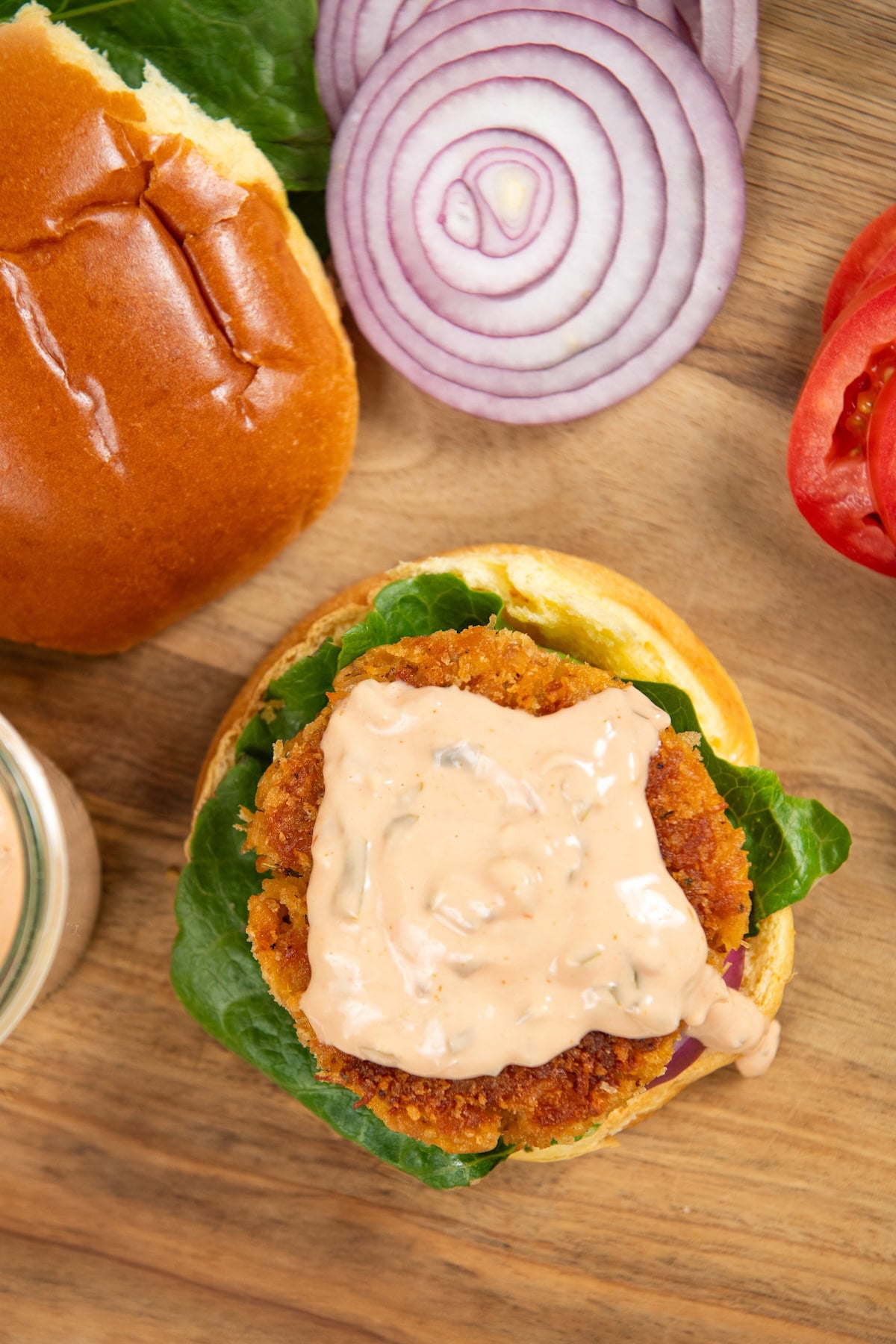 This Chicken Patty Sandwich turns two cans of chicken into a quick and easy dinner for six! Served with a tangy sauce and all the toppings. Chicken patty sandwich with lettuce, and creamy sauce on a bun, next to sliced red onion, tomato, and a top bun, on a wooden surface.