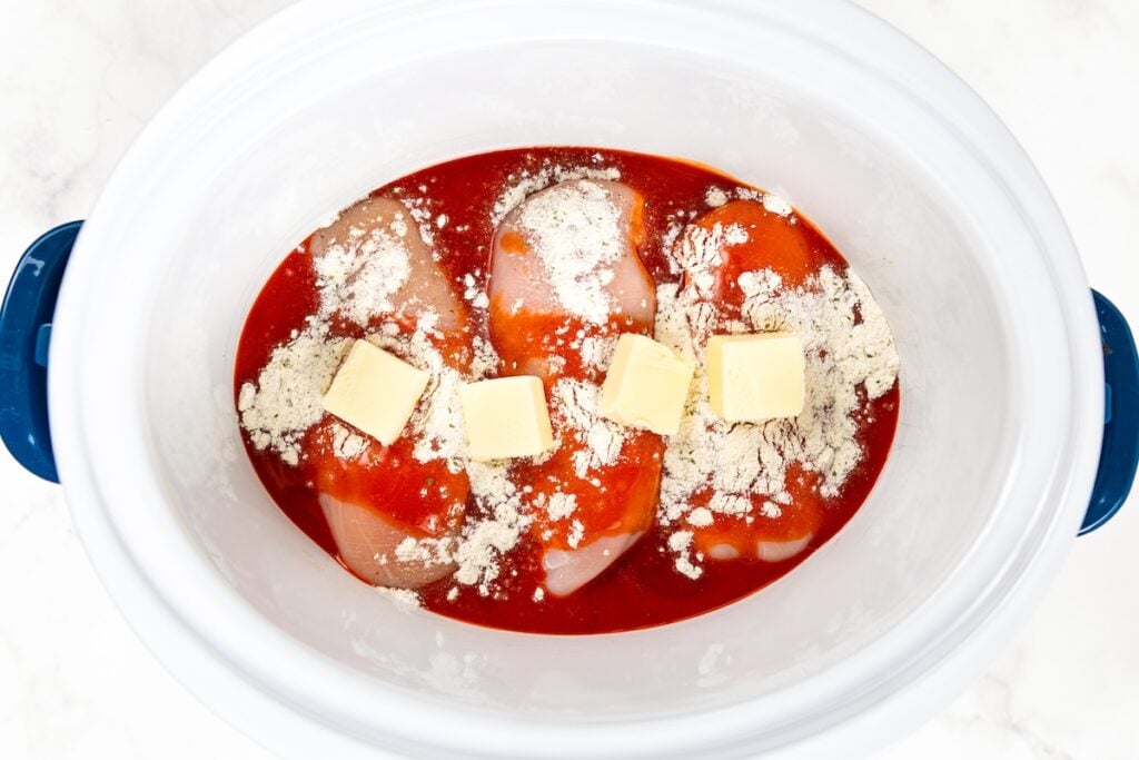 Crockpot Buffalo Chicken is creamy, tangy, a little spicy, and made with just 6 ingredients. Dump it in, shred it, and dinner is ready! Raw chicken breasts in a slow cooker with buffalo sauce, dry ranch seasoning mix, and three pats of butter on top, all ready to be cooked.