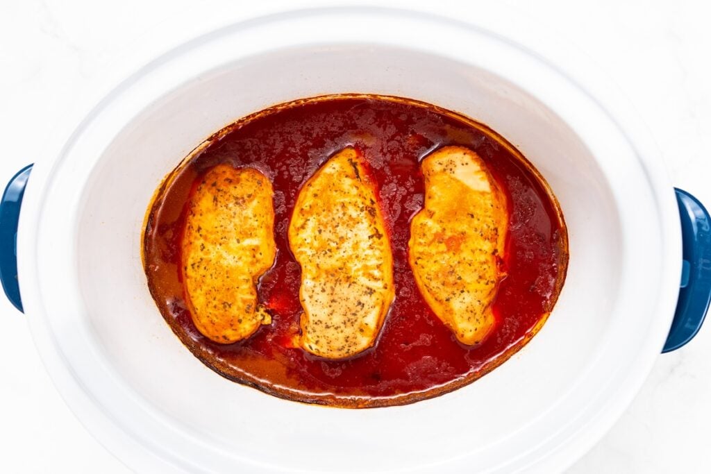 Crockpot Buffalo Chicken is creamy, tangy, a little spicy, and made with just 6 ingredients. Dump it in, shred it, and dinner is ready! Three seasoned chicken breasts cooked in a buffalo sauce inside a white crockpot with blue handles, shown from above.
