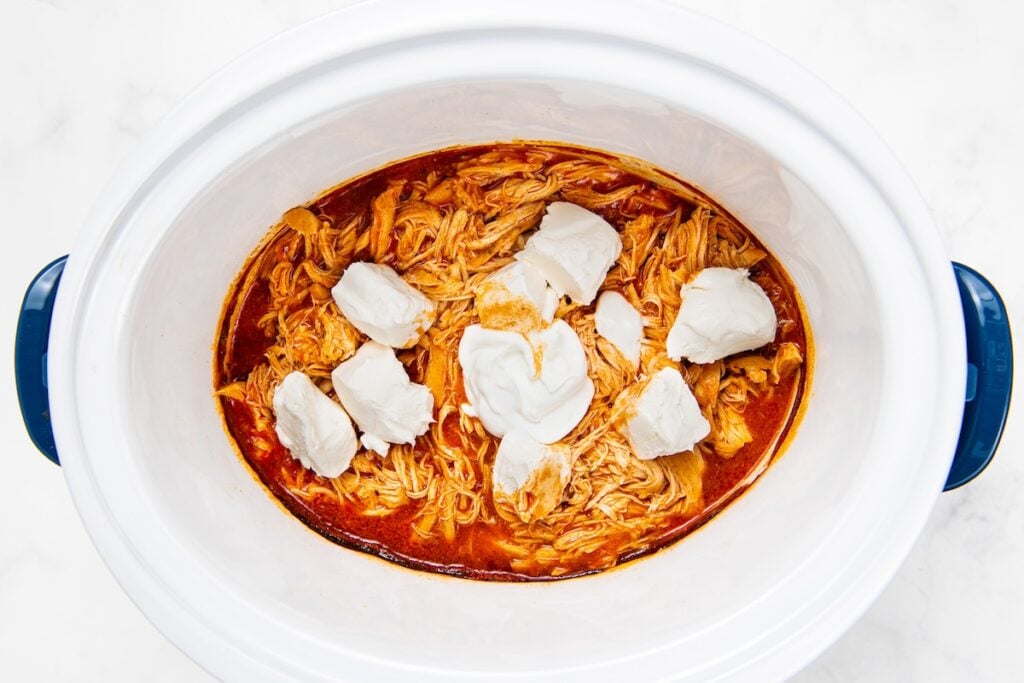 Crockpot Buffalo Chicken is creamy, tangy, a little spicy, and made with just 6 ingredients. Dump it in, shred it, and dinner is ready! Shredded slow cooker buffalo chicken in red sauce inside a white crockpot, topped with several dollops of cream cheese.
