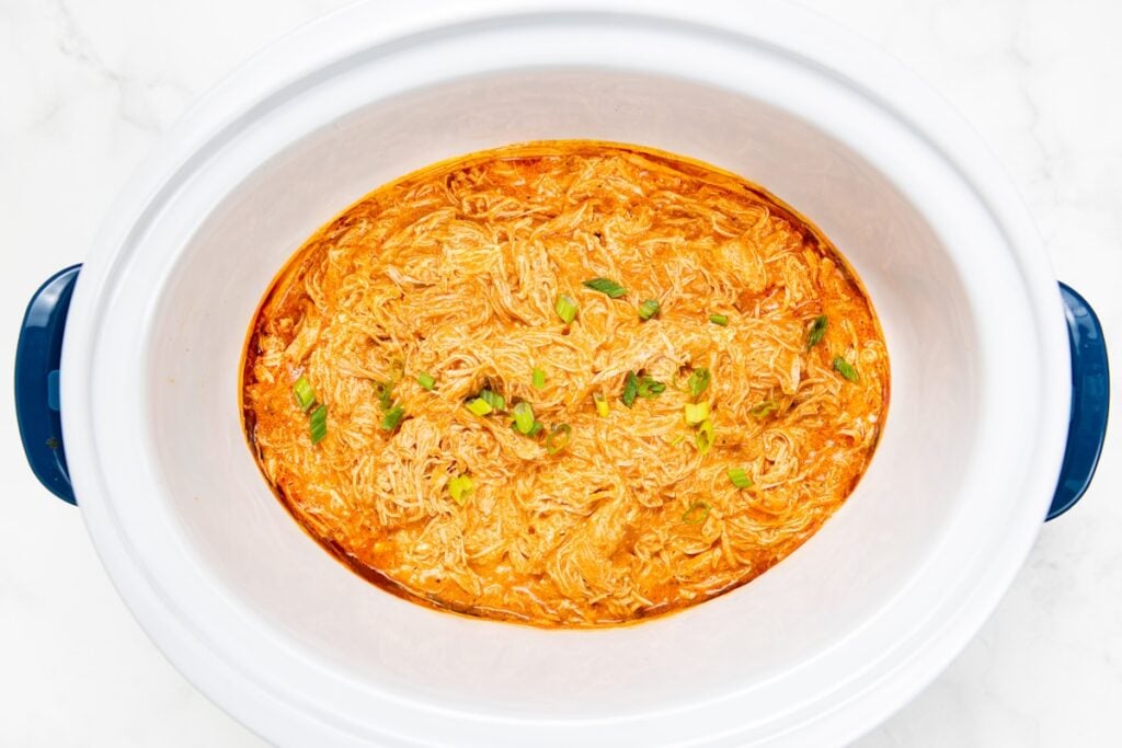 Crockpot Buffalo Chicken is creamy, tangy, a little spicy, and made with just 6 ingredients. Dump it in, shred it, and dinner is ready! Shredded crockpot buffalo chicken in a rich orange sauce garnished with fresh herbs, served in a white slow cooker with blue handles on a white background.
