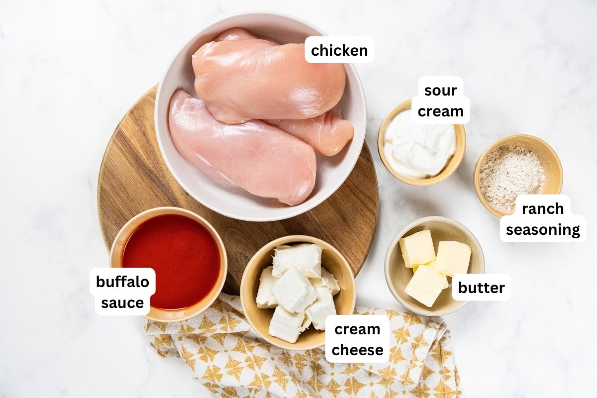 Crockpot Buffalo Chicken is creamy, tangy, a little spicy, and made with just 6 ingredients. Dump it in, shred it, and dinner is ready! Raw chicken breasts in a bowl on a cutting board, surrounded by labeled bowls of buffalo sauce, cream cheese, sour cream, ranch seasoning, and butter on a white surface.