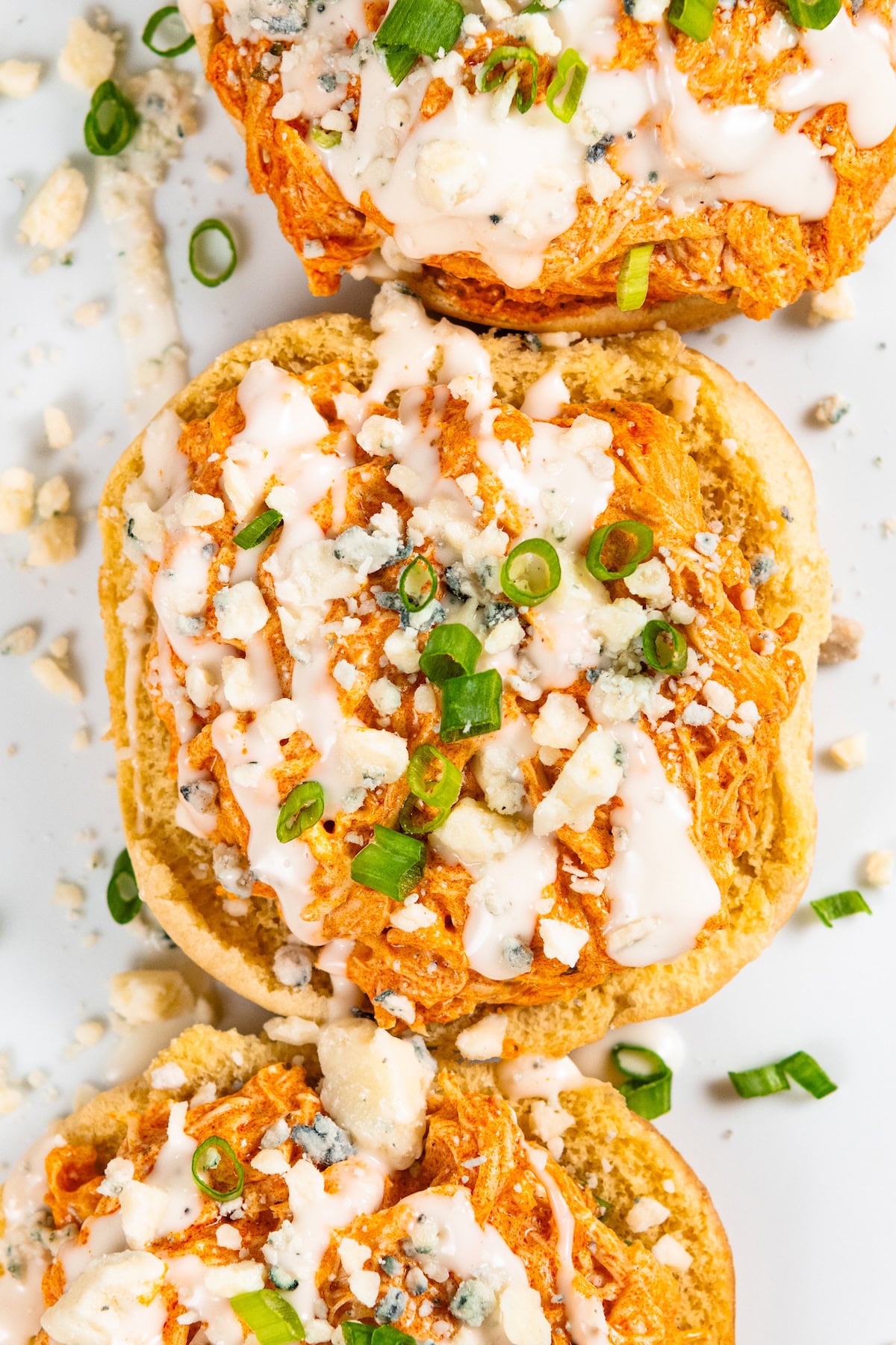 Crockpot Buffalo Chicken is creamy, tangy, a little spicy, and made with just 6 ingredients. Dump it in, shred it, and dinner is ready! Three sandwich buns topped with crockpot buffalo chicken, blue cheese crumbles, chopped green onions, and a drizzle of ranch, arranged on a white surface.
