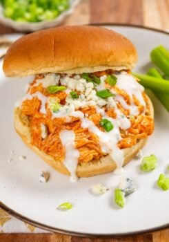 A crockpot buffalo chicken sandwich, topped with ranch dressing, blue cheese crumbles, and chopped green onions, served on a white plate with celery sticks on the side.