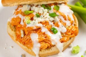 A crockpot buffalo chicken sandwich, topped with ranch dressing, blue cheese crumbles, and chopped green onions, served on a white plate with celery sticks on the side.