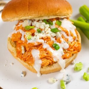 A crockpot buffalo chicken sandwich, topped with ranch dressing, blue cheese crumbles, and chopped green onions, served on a white plate with celery sticks on the side.