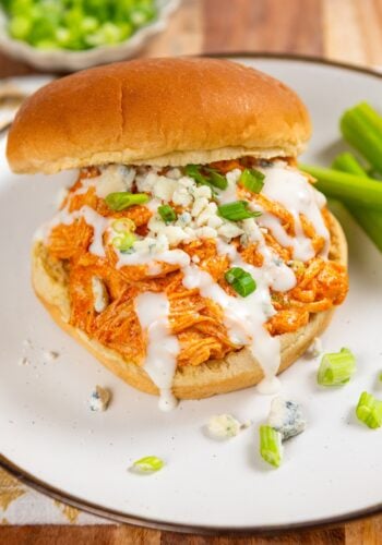 A crockpot buffalo chicken sandwich, topped with ranch dressing, blue cheese crumbles, and chopped green onions, served on a white plate with celery sticks on the side.