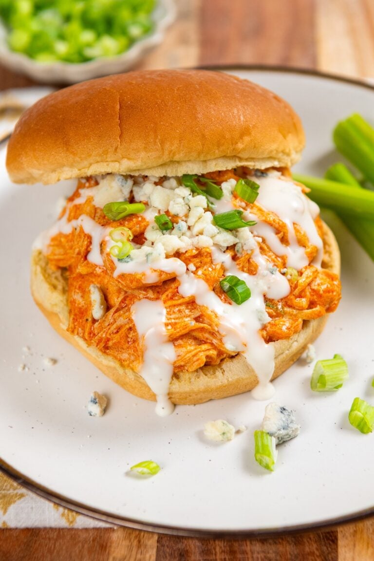 A crockpot buffalo chicken sandwich, topped with ranch dressing, blue cheese crumbles, and chopped green onions, served on a white plate with celery sticks on the side.