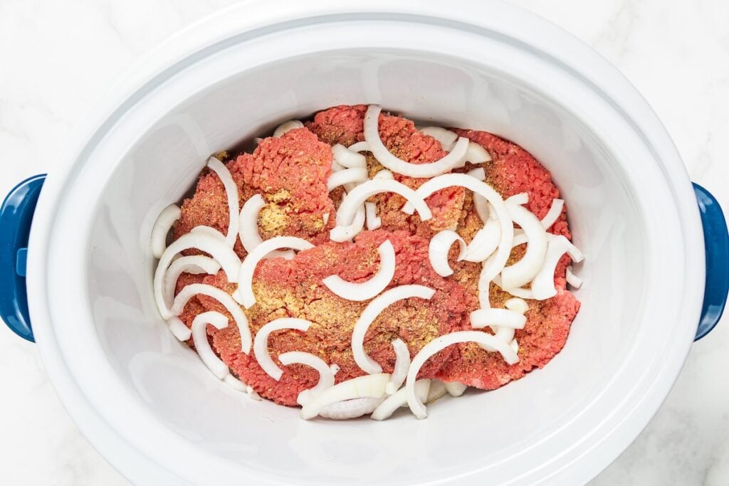 Crockpot Cube Steak is smothered in a rich and thick brown gravy with onions until it's fall-apart tender. Set it and forget it! Raw beef, sliced onions, and seasonings in a white slow cooker with blue handles, ready to be cooked.