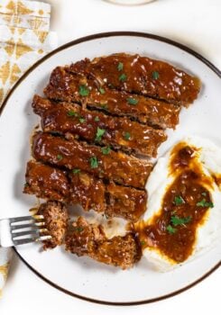Sliced crockpot cube steak and gravy is served next to mashed potatoes with gravy on a white plate.