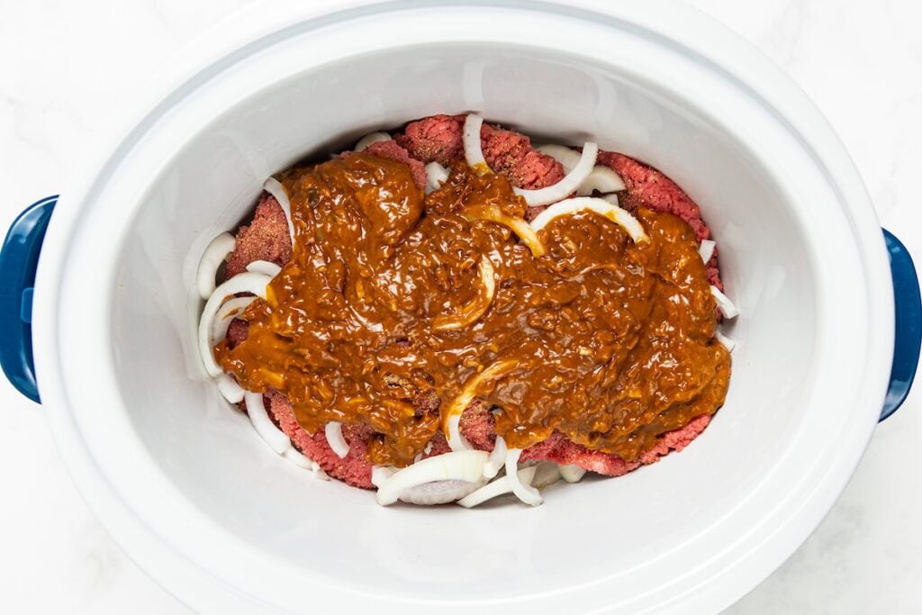 Crockpot Cube Steak is smothered in a rich and thick brown gravy with onions until it's fall-apart tender. Set it and forget it! A slow cooker filled with raw cube steak, sliced onions, and a layer of gravy mix on top, ready to be cooked.