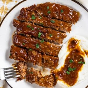 Sliced crockpot cube steak and gravy is served next to mashed potatoes with gravy on a white plate.