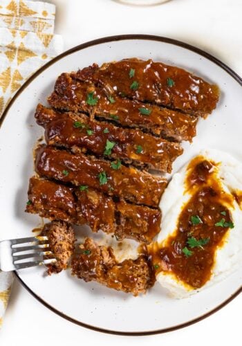 Sliced crockpot cube steak and gravy is served next to mashed potatoes with gravy on a white plate.