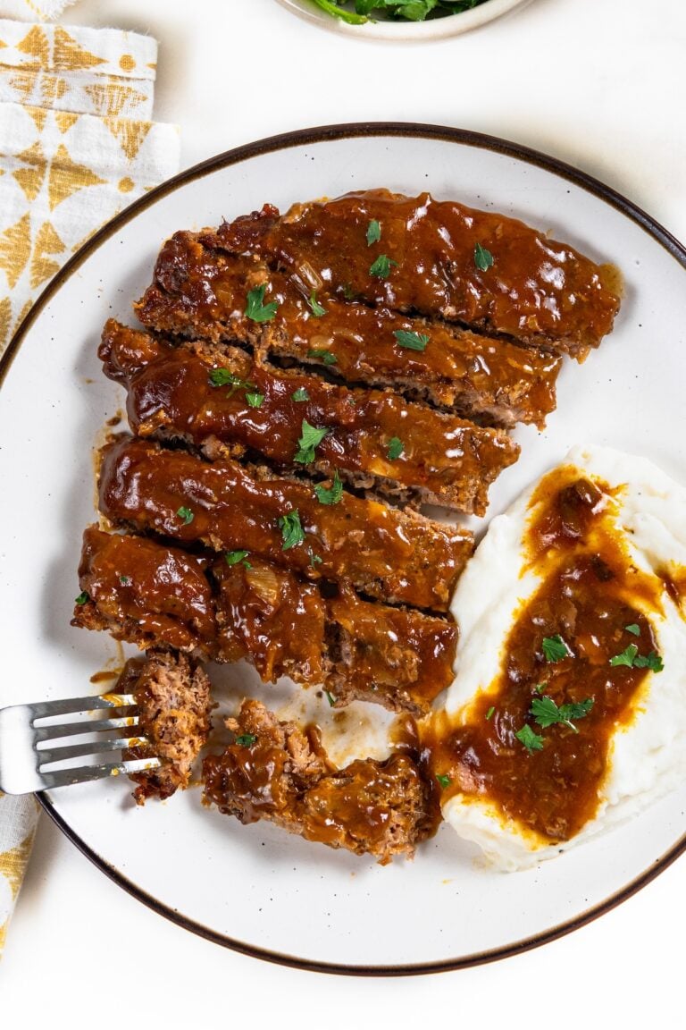 Sliced crockpot cube steak and gravy is served next to mashed potatoes with gravy on a white plate.