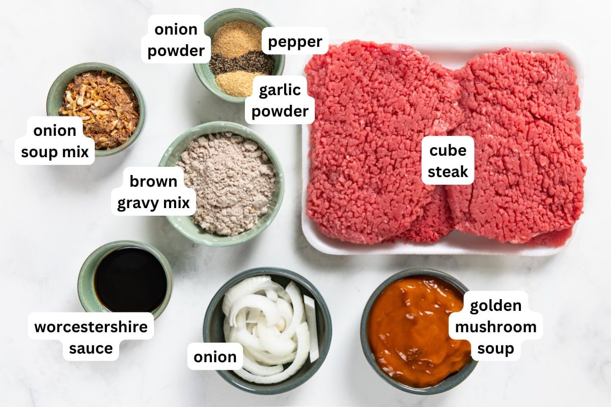 Crockpot Cube Steak is smothered in a rich and thick brown gravy with onions until it's fall-apart tender. Set it and forget it! Ingredients are laid out on a white surface, each labeled: cube steak, brown gravy mix, onion soup mix, onion powder, pepper, garlic powder, Worcestershire sauce, onion, and golden mushroom soup.