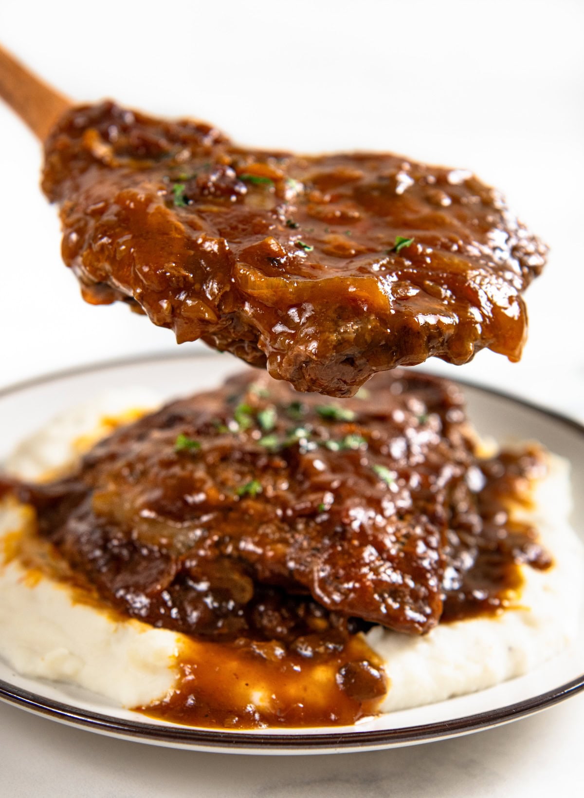 Crockpot Cube Steak is smothered in a rich and thick brown gravy with onions until it's fall-apart tender. Set it and forget it! A wooden spoon lifts a piece of smothered crockpot cube steak and gravy over a plate of mashed potatoes topped with more cube steak and sauce.