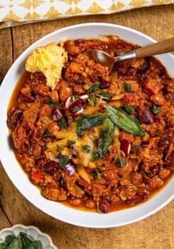 A bowl of crockpot pumpkin turkey chili topped with melted cheese, crispy sage, and diced red onions. A piece of cornbread with butter is on the side of the bowl, and a spoon is partially in the chili. The bowl sits on a wooden surface.