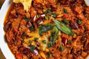 A bowl of crockpot pumpkin turkey chili topped with melted cheese, crispy sage, and diced red onions. A piece of cornbread with butter is on the side of the bowl, and a spoon is partially in the chili. The bowl sits on a wooden surface.
