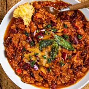 A bowl of crockpot pumpkin turkey chili topped with melted cheese, crispy sage, and diced red onions. A piece of cornbread with butter is on the side of the bowl, and a spoon is partially in the chili. The bowl sits on a wooden surface.