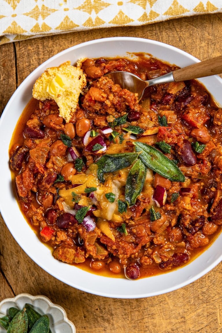 A bowl of crockpot pumpkin turkey chili topped with melted cheese, crispy sage, and diced red onions. A piece of cornbread with butter is on the side of the bowl, and a spoon is partially in the chili. The bowl sits on a wooden surface.